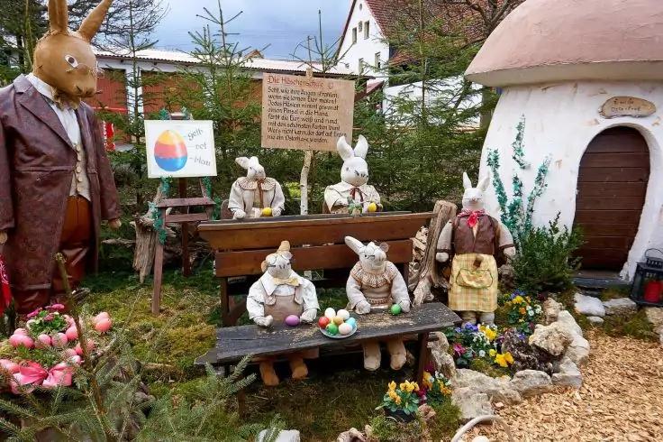 Mehrere Hasenfiguren aus Holz, einige sitzen an Schulb&auml;nken mit bemalten Eiern, ein gro&szlig;er Hase im Mantel links, ein rundes H&auml;uschen rechts, Schilder, Tannenb&auml;ume und Blumen im Freien