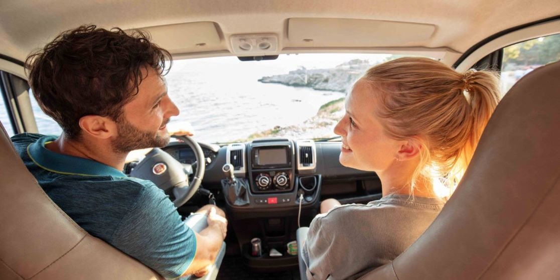 Mann und Frau sitzen in den vorderen Sitzen eines Fahrzeugs, blicken zueinander; Armaturenbrett und Lenkrad sichtbar; durch die Frontscheibe Meer und Felsen