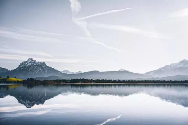 Spiegelglatter See vor einer Bergkette mit schneebedeckten Gipfeln, Baumreihe am Ufer, blasser Himmel mit Kondensstreifen, Spiegelung im Wasser
