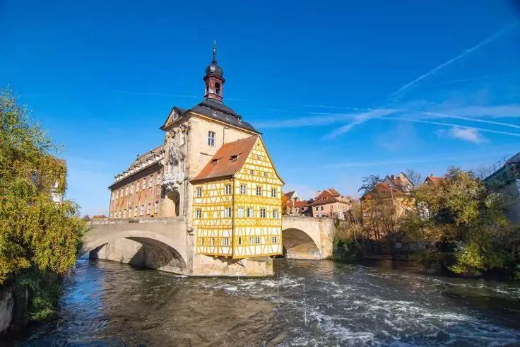 Gelbes Fachwerkgeb&auml;ude mit kleinem Turm auf einer Steinbr&uuml;cke &uuml;ber einem flie&szlig;enden Fluss; Ufer mit B&auml;umen und Geb&auml;uden, blauer Himmel mit Kondensstreifen