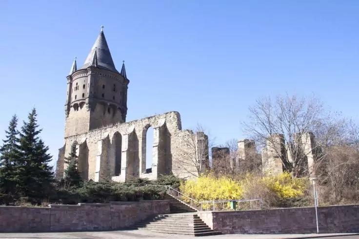 Steinerner Turm mit spitzem Dach neben steinernen Ruinen mit Bogen&ouml;ffnungen; im Vordergrund Stufen, immergr&uuml;ne und kahle B&auml;ume sowie gelb bl&uuml;hende Str&auml;ucher; blauer Himmel