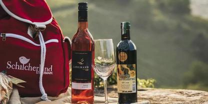 Zwei Weinflaschen, ein leeres Weinglas, roter Rucksack und Landschaft im Hintergrund