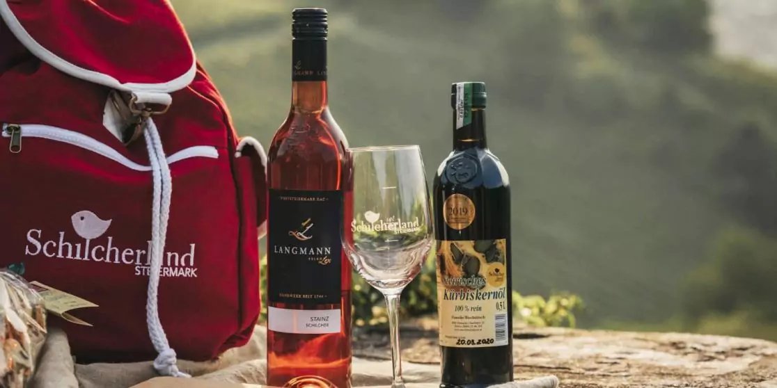 Zwei Weinflaschen, ein leeres Weinglas, roter Rucksack und Landschaft im Hintergrund