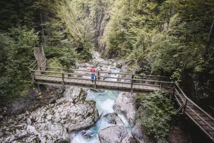 Holzsteg &uuml;ber einem t&uuml;rkisfarbenen, felsigen Bach in einer schmalen, bewaldeten Schlucht; zwei Personen in roten und blauen Jacken auf dem Steg; Holztreppe rechts