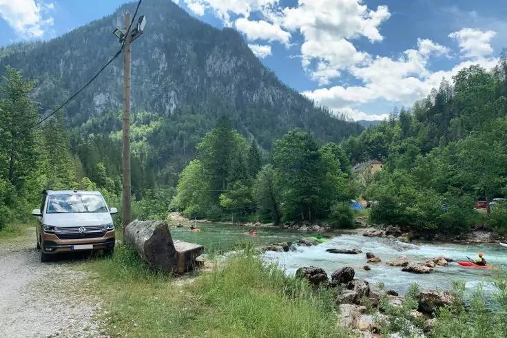 Van steht auf einem Schotterweg links, Holzbank neben gro&szlig;em Felsen, Fluss mit Felsen und rotem Kajak rechts, B&auml;ume und Berg im Hintergrund, bew&ouml;lkter Himmel