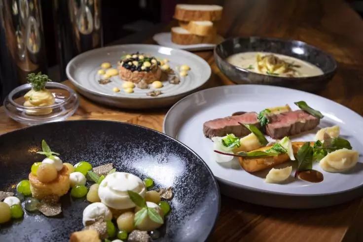 Mehrere Teller mit Speisen auf einem Holztisch: schwarzer Teller mit wei&szlig;er Creme, kleinen gr&uuml;nen Kugeln und Kr&uuml;meln; wei&szlig;er Teller mit rosa Fleischscheiben, Kartoffelst&uuml;cken, hellem P&uuml;ree und gr&uuml;nen Bl&auml;ttern; Schale mit heller Suppe und Brotscheiben