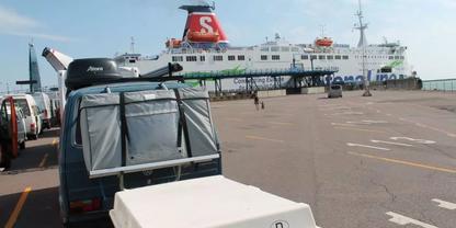 F&auml;hre mit rotem Schornstein und wei&szlig;em 'S' am Kai, im Vordergrund Gep&auml;ckwagen und wei&szlig;er Container, wenige Personen auf dem Hafenbereich, klarer Himmel