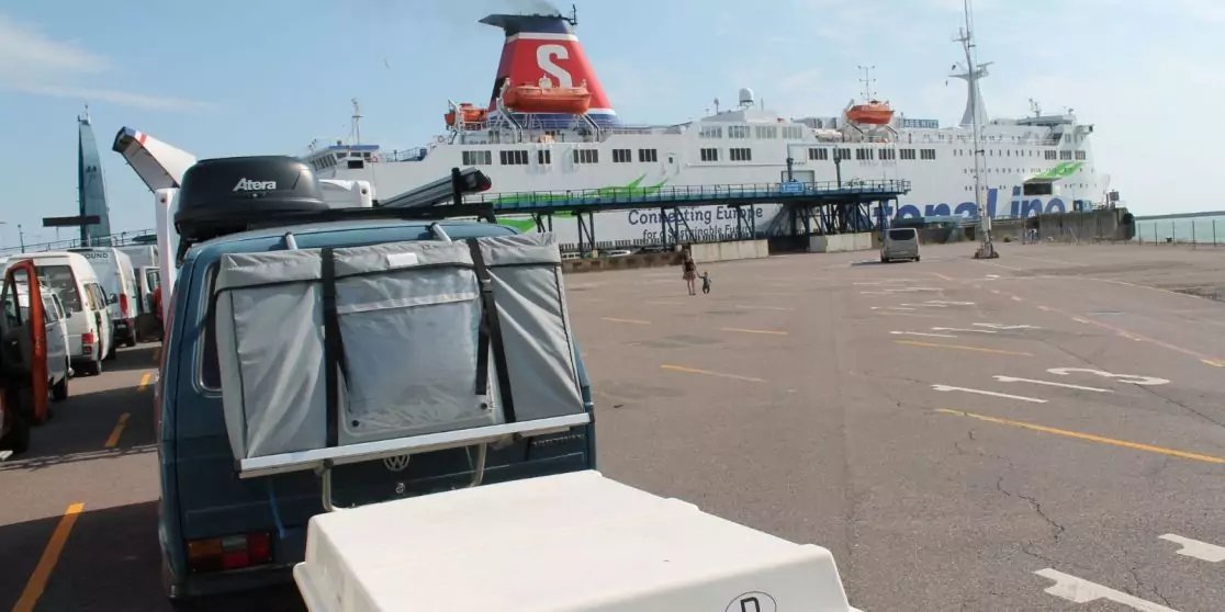 F&auml;hre mit rotem Schornstein und wei&szlig;em 'S' am Kai, im Vordergrund Gep&auml;ckwagen und wei&szlig;er Container, wenige Personen auf dem Hafenbereich, klarer Himmel