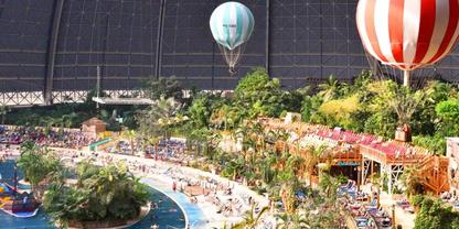 Gro&szlig;e &uuml;berdachte Halle mit gew&ouml;lbtem Glasdach, tropischen Pflanzen und Palmen, zwei Hei&szlig;luftballons, langes Poolbecken mit Liegen und Wegen, Reihen bunter Geb&auml;ude und Menschen