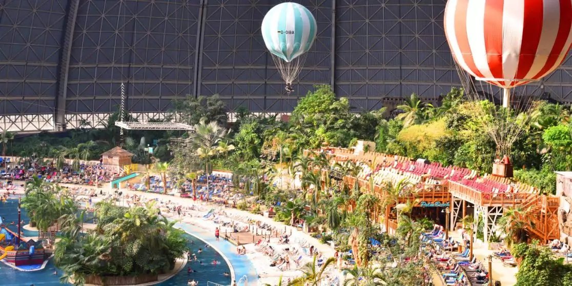 Gro&szlig;e &uuml;berdachte Halle mit gew&ouml;lbtem Glasdach, tropischen Pflanzen und Palmen, zwei Hei&szlig;luftballons, langes Poolbecken mit Liegen und Wegen, Reihen bunter Geb&auml;ude und Menschen