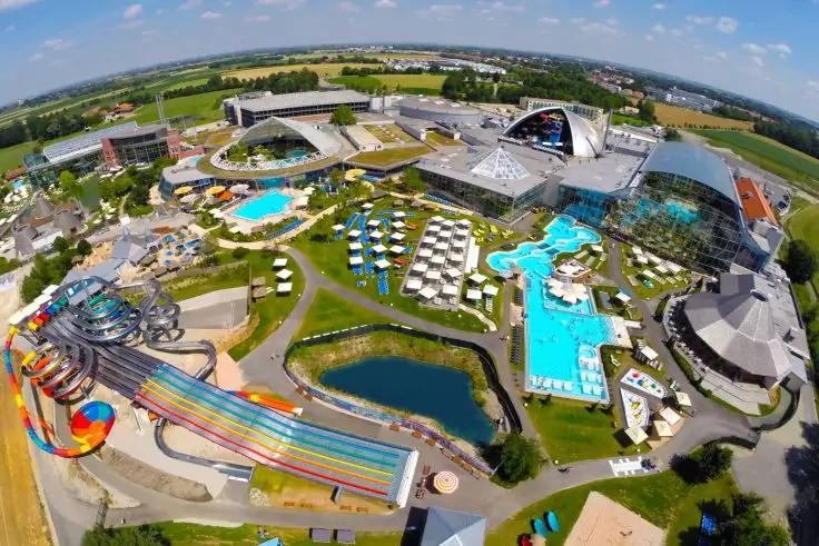 Luftaufnahme mit mehreren Au&szlig;enpools, bunten Wasserrutschen, Liegen, Sonnenschirmen, einem Teich und umliegenden Geb&auml;uden