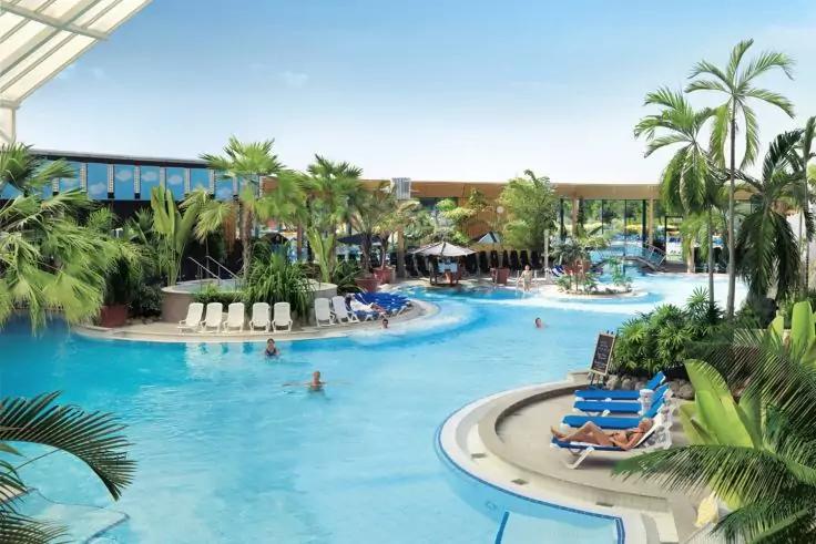 Gro&szlig;er geschwungener Au&szlig;enpool mit blauem Wasser, Palmen und Pflanzen, Sonnenliegen am Beckenrand, Sonnenschirme, einzelne Personen im Wasser und Hotelgeb&auml;ude im Hintergrund