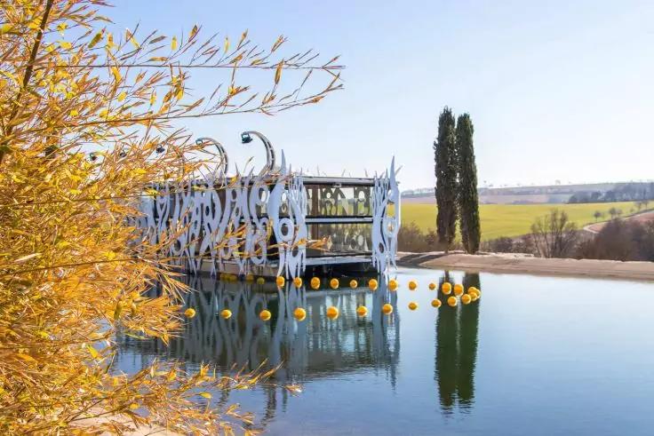 Wei&szlig;es ornamentales Pavillon mit ausgeschnittenen Mustern auf einem spiegelnden Wasserbecken, Reihe gelber Bojen, gelbe Str&auml;ucher links, zwei hohe Zypressen im Hintergrund, weiter Feldhorizont unter blauem Himmel