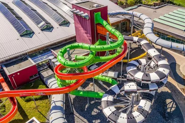 Luftaufnahme mehrerer geschwungener, bunter Wasserrutschen (gr&uuml;n, rot, gelb) &uuml;ber Geb&auml;uded&auml;chern mit Solarpaneelen