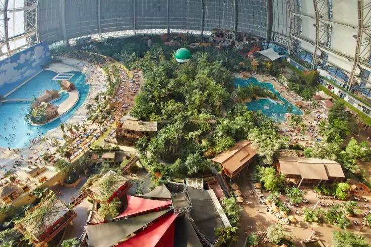 Innenraum unter einer Glasdachkuppel mit zentraler dicht bewachsener Insel, mehreren Pools und Sandfl&auml;chen, Wegen, Holzh&auml;usern und vielen Personen auf Liegest&uuml;hlen