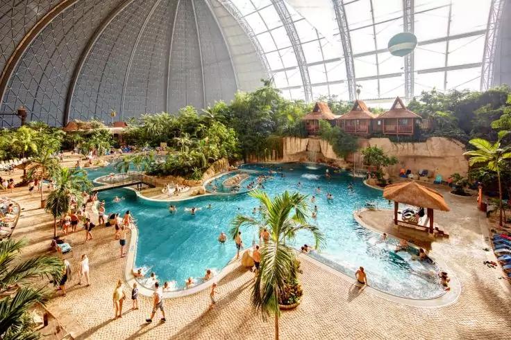 Gro&szlig;e Halle mit gebogenem Glasdach, mehrere verbundene Wasserbecken, Insel mit Palmen, strohgedeckte H&uuml;tten an einer Felswand, Treppen und Liegest&uuml;hle, Menschen in Badekleidung