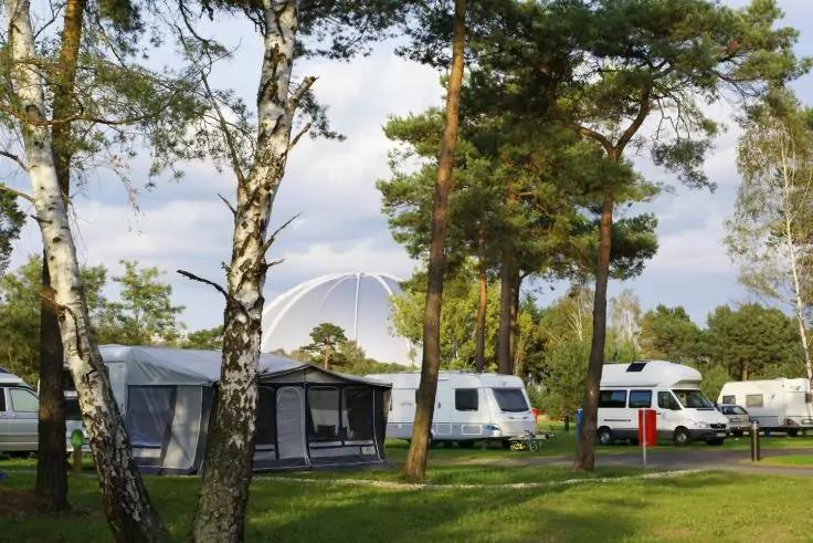 Mehrere Wohnwagen und Wohnmobile auf einem Campingplatz zwischen Birken und Kiefern, wei&szlig;e Kuppel im Hintergrund, Grasfl&auml;chen