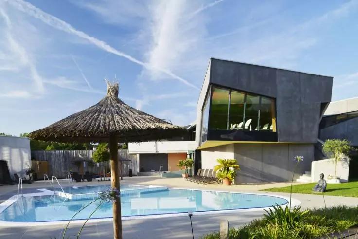 Schwimmbecken im Freien mit Strohsonnenschirm, Einstiegsleiter, Pflanzen und Geb&auml;ude mit gro&szlig;en Fenstern unter blauem Himmel mit Kondensstreifen