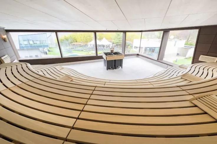Gebogene, gestufte Holzsitze vor gro&szlig;en Panoramafenstern; mittig ein kleiner Tisch mit einem Stuhl; drau&szlig;en Wasser, Boote und Geb&auml;ude sichtbar