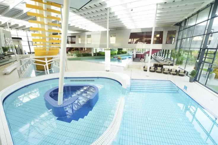 Hallenbad mit rechteckigem Becken, blauem Wasser, zentraler blauer spiralf&ouml;rmiger Konstruktion mit Treppenstufen, gro&szlig;en Panoramafenstern und Sitzbereichen