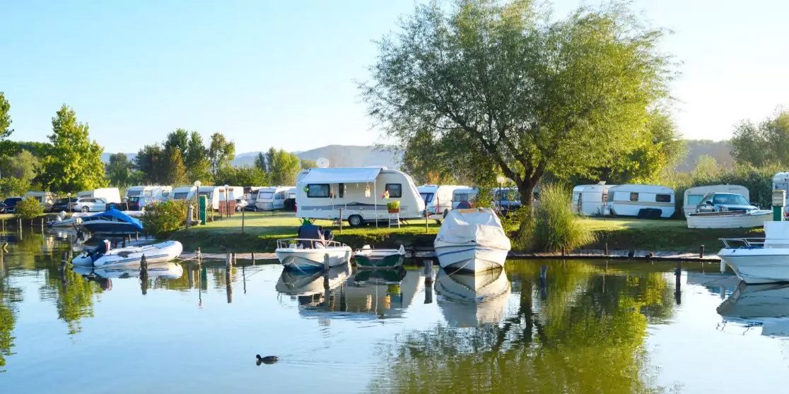 Mehrere Boote an einem Steg, Wohnwagen am Ufer, gro&szlig;er Baum, Ente und Spiegelungen im ruhigen Wasser, blauer Himmel