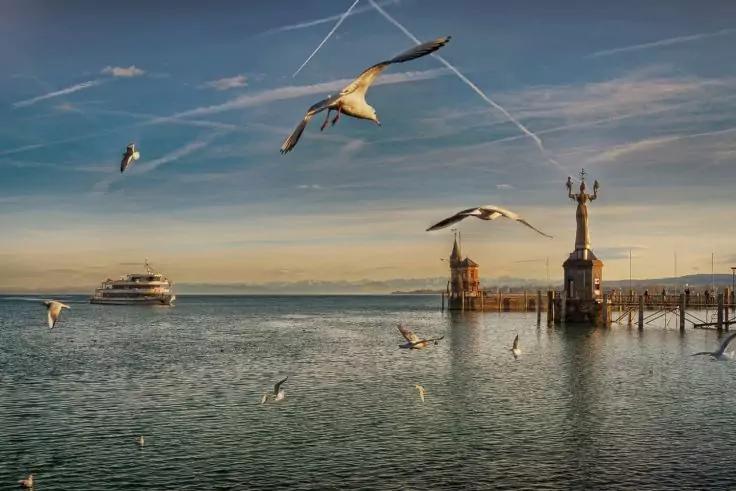 Wasserfl&auml;che mit fliegenden M&ouml;wen, Steg rechts mit hoher Turmskulptur, F&auml;hre links am Horizont, Himmel mit Kondensstreifen