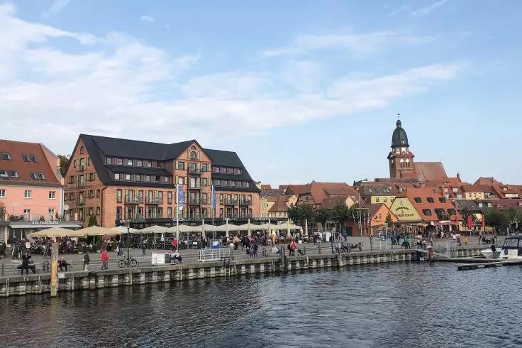 Uferpromenade mit Menschen, historischen H&auml;usern und einem Turm, Steg und ruhigem Wasser unter teilweise bew&ouml;lktem Himmel