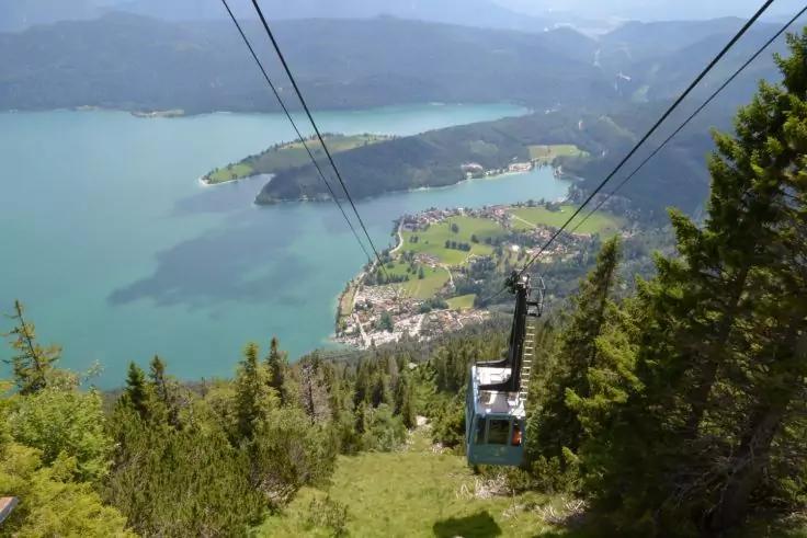 Seilbahnkorb an zwei Stahlseilen &uuml;ber einem bewaldeten Hang, darunter ein t&uuml;rkisfarbener See mit Inseln und Uferort, im Hintergrund Berge