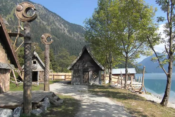 Uferweg mit kleinen Holzh&auml;usern, geschnitzten Holzpfosten, B&auml;umen und einem Boot am klaren Bergsee, Berge im Hintergrund