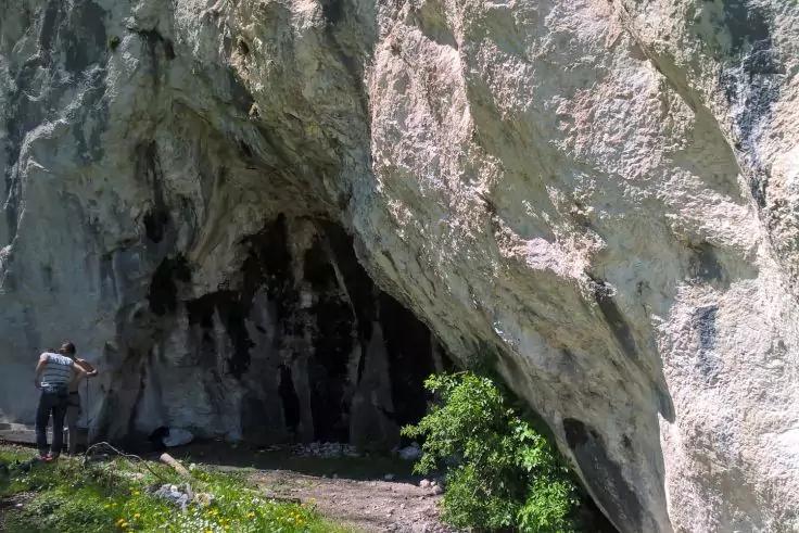Gro&szlig;e H&ouml;hlen&ouml;ffnung unter einem Fels&uuml;berhang; links steht eine Person; im Vordergrund Gras und ein Weg