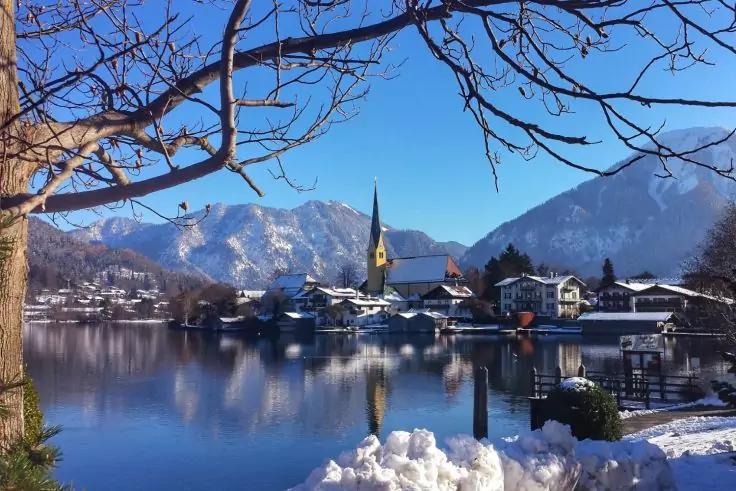 See mit Spiegelung, Kirche mit spitzem Turm am Ufer, H&auml;user, Schnee an Ufer und auf D&auml;chern, Bergkette im Hintergrund, kahle Baum&auml;ste im Vordergrund, blauer Himmel