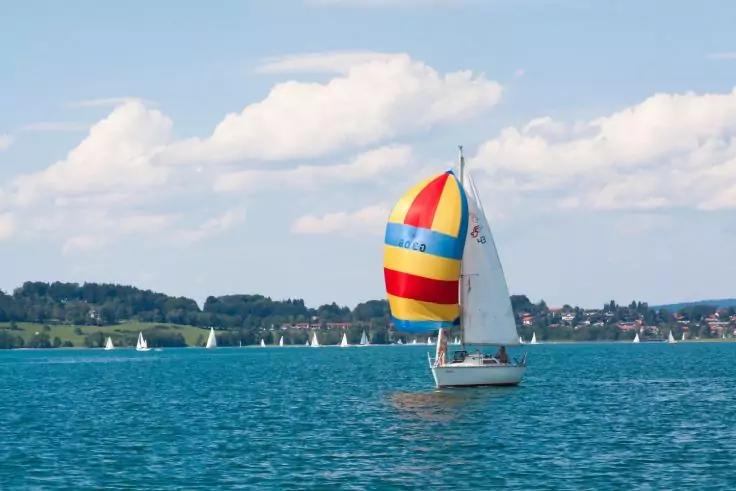 Segelboot mit rot-gelb-blau gestreiftem Spinnaker auf blauem See, mehrere wei&szlig;e Segelboote in der Ferne, Ufer mit B&auml;umen und H&auml;usern, wolkiger Himmel