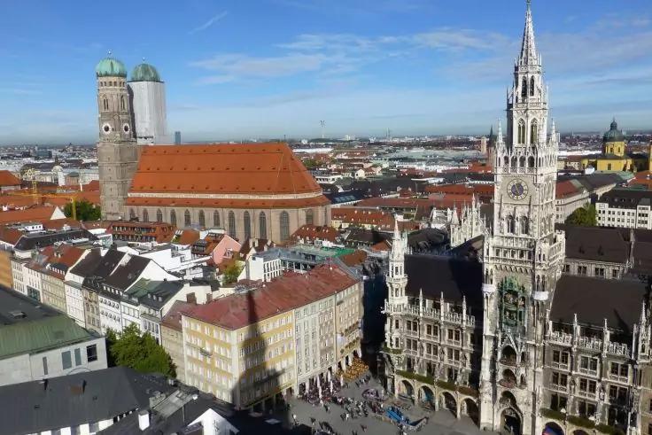 Vogelperspektive auf einen Platz mit links einer gro&szlig;en Kirche mit zwei gr&uuml;nen Kuppeln, rechts einem hohen verzierten Turm, umliegenden mehrst&ouml;ckigen Geb&auml;uden und Menschen auf dem Platz