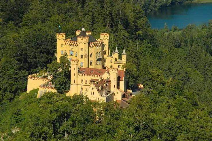 Gelbes Schloss mit mehreren T&uuml;rmen, umgeben von dichtem Wald, See im Hintergrund