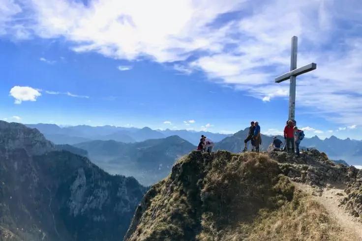 Berggipfel mit gro&szlig;em Kreuz, mehreren Personen auf schmalem Grat und Bergpanorama unter blauem Himmel mit Wolken