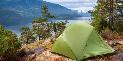 Gr&uuml;nes Zelt auf felsiger Uferb&ouml;schung, Nadelb&auml;ume, See und Berge im Hintergrund, teils bew&ouml;lkter Himmel