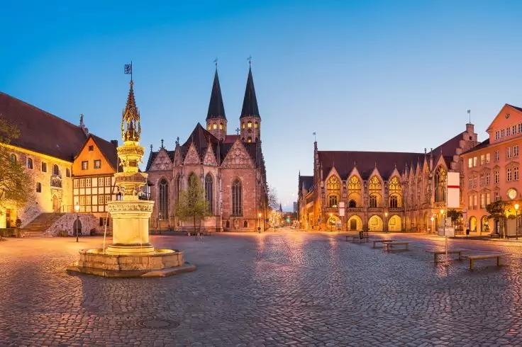 Gepflasterter Marktplatz bei Dämmerung mit zentralem Brunnen und Statue, Kirche mit zwei Spitztürmen, umgebende Fachwerk- und Steingebäude, Straßenlaternen, keine Personen