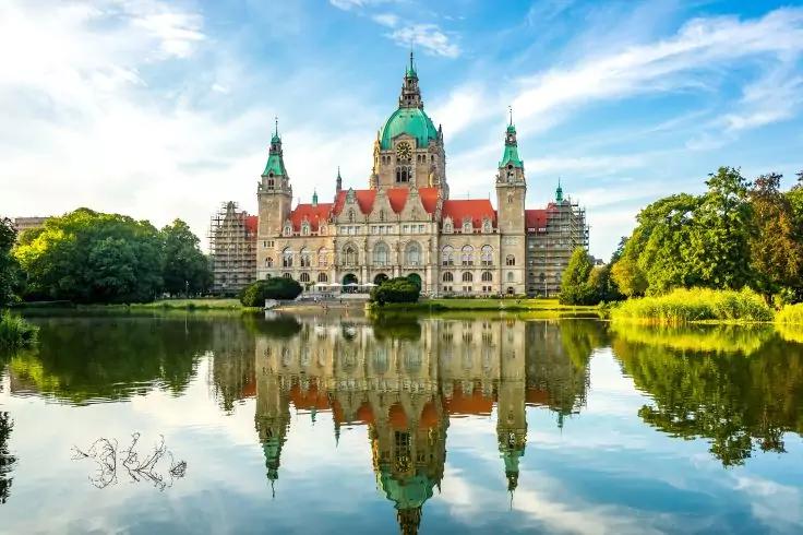 Historisches Gebäude mit grüner Kuppel und mehreren Türmen, Fassade spiegelt sich in einem Teich, umgeben von Bäumen, blauer Himmel mit Wolken