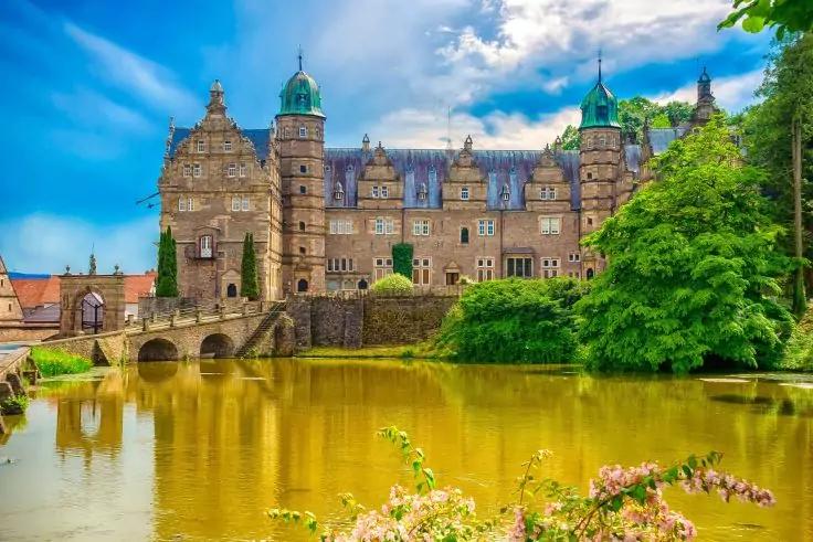 Großes steinernes Gebäude mit mehreren Türmen und Zinnen, davor eine Bogenbrücke über einen Teich, spiegelndes Wasser, Bäume und bewölkter Himmel