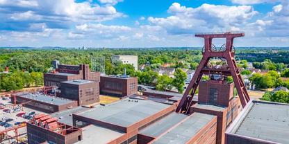 Luftaufnahme eines Industriekomplexes mit flachen roten Backstein- und Metallgeb&auml;uden, einem hohen roten Stahlger&uuml;st-Turm mit rechteckiger Plattform, B&auml;umen und blauem Himmel mit wei&szlig;en Wolken