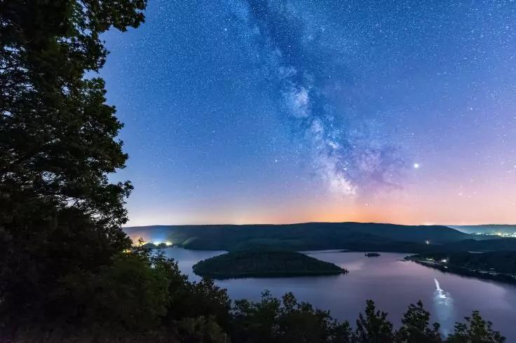 Sternenhimmel mit sichtbarer Milchstra&szlig;e &uuml;ber einem See mit Inseln, dunklen H&uuml;geln am Horizont, Uferlichtern und B&auml;umen im Vordergrund