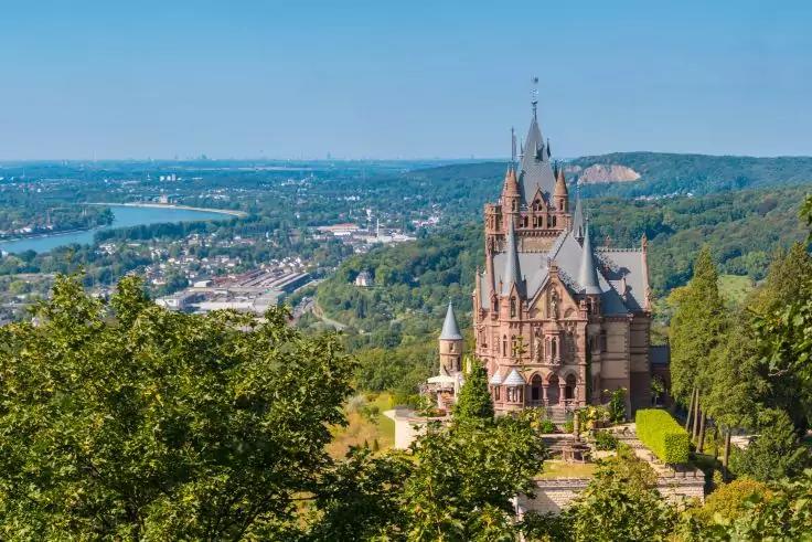 Gro&szlig;es steinernes Schloss auf einem bewaldeten H&uuml;gel, darunter Stadt und Fluss im Tal, blauer Himmel