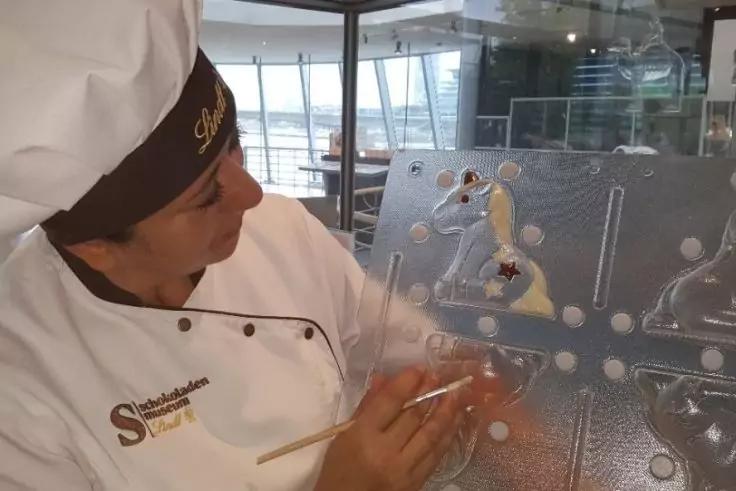 Person in wei&szlig;er Jacke und braunem Hut h&auml;lt einen d&uuml;nnen Pinsel vor einer gl&auml;nzenden, durchsichtigen Platte mit geformten Dekorationen; Fenster im Hintergrund