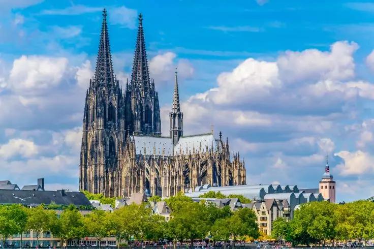 Gro&szlig;es steinernes Kirchengeb&auml;ude mit zwei hohen T&uuml;rmen am Flussufer; im Vordergrund gr&uuml;ne B&auml;ume und niedrige H&auml;user; blauer Himmel mit wei&szlig;en Wolken