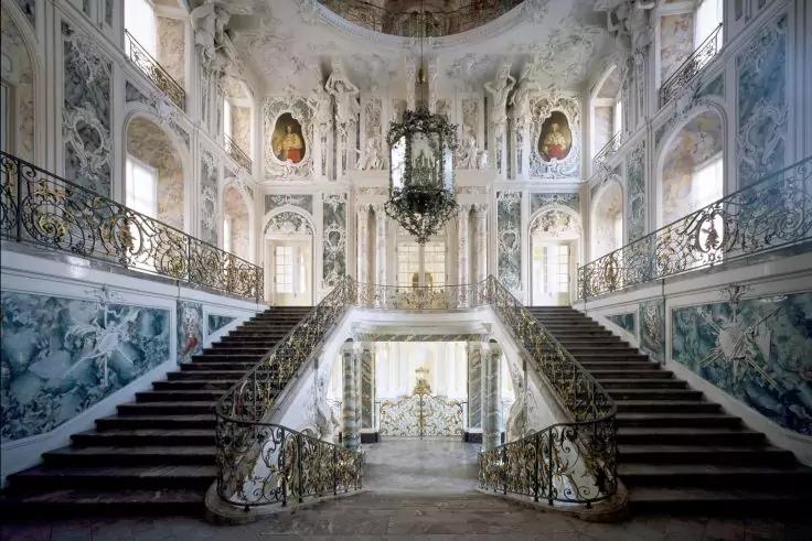 Breite geschwungene Doppeltreppe mit schmiedeeisernen Gel&auml;ndern, h&auml;ngender Kronleuchter, verzierte Stuckw&auml;nde, bemalte Decke und gro&szlig;e Fenster