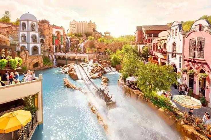 Boot f&auml;hrt eine Abfahrt in einem Wasserkanal zwischen bunten Geb&auml;uden, gro&szlig;e Wasserspritzer, Menschen auf Balkonen und Promenade, B&auml;ume und Sonnenschein