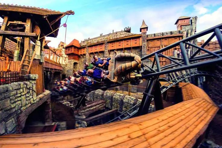 Achterbahnzug mit sitzenden Personen auf gebogener Stahlstrecke vor h&ouml;lzernen Geb&auml;uden mit T&uuml;rmen und steinernen Mauern, blauer Himmel