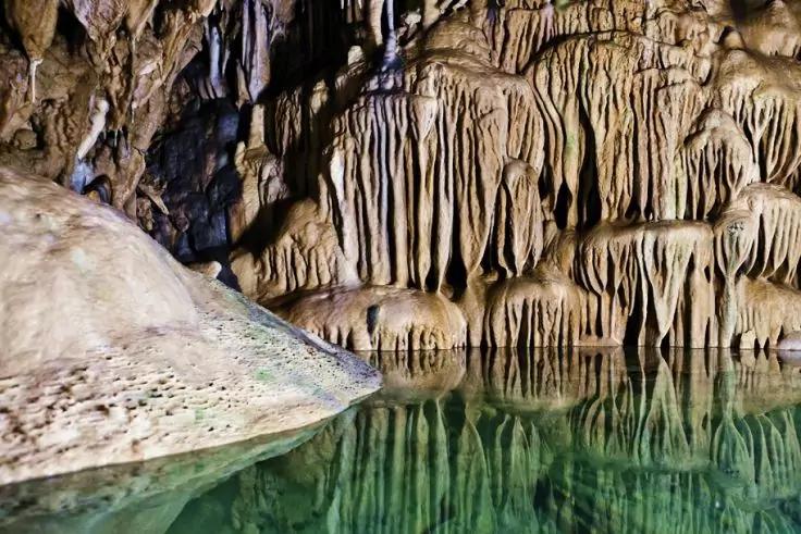 H&ouml;hleninnenraum mit Tropfsteinformationen an der Decke, felsiger Vordergrund links und gr&uuml;nlich reflektierendes stehendes Wasser