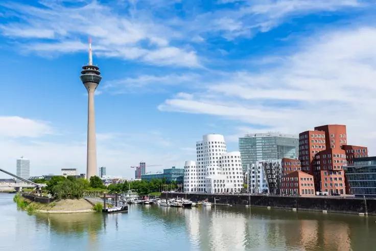 Flussufer mit spiegelndem Wasser, mehrere Boote, moderne Geb&auml;ude (wei&szlig;e geschwungene, rote Backstein- und Glasfassaden) und ein hoher Turm mit runder Plattform unter blauem Himmel mit Wolken