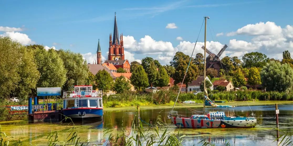 Boote auf einem Fluss, im Hintergrund eine Backsteinkirche mit zwei spitzen T&uuml;rmen, eine Windm&uuml;hle, B&auml;ume und blauer Himmel mit Wolken